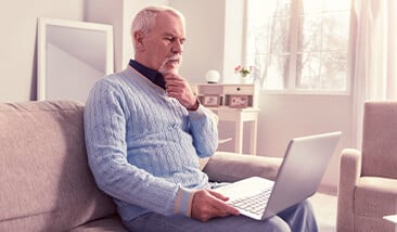 Older gentleman in light blue sweater looking at his laptop contemplating cremation decision