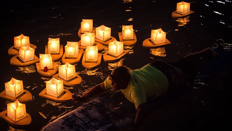 memorial service candles floating on water at night, with man on surfboard guiding them