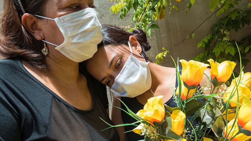 mother and daugher wearing covid masks and holding yellow flowers at outdoor funeral in cemetery in LA