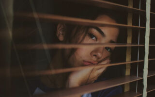 young woman looking through window blinds while processing grief in her santa monica home