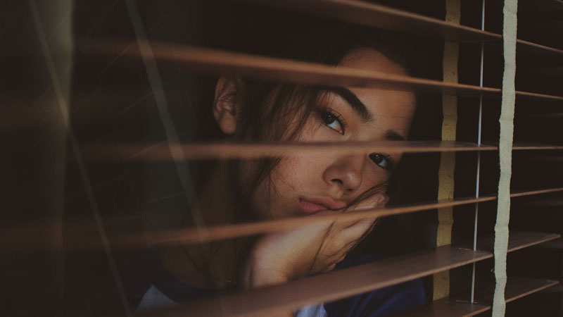 young woman looking through window blinds while processing grief in her santa monica home